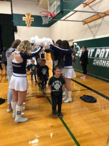 Elementary students celebrate homecoming with high school cheerleaders in a gym.