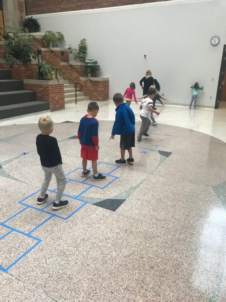 A group of children walk along tape on the floor.