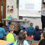 School Counselor standing at the smart board while group of children sit on the floor listening.