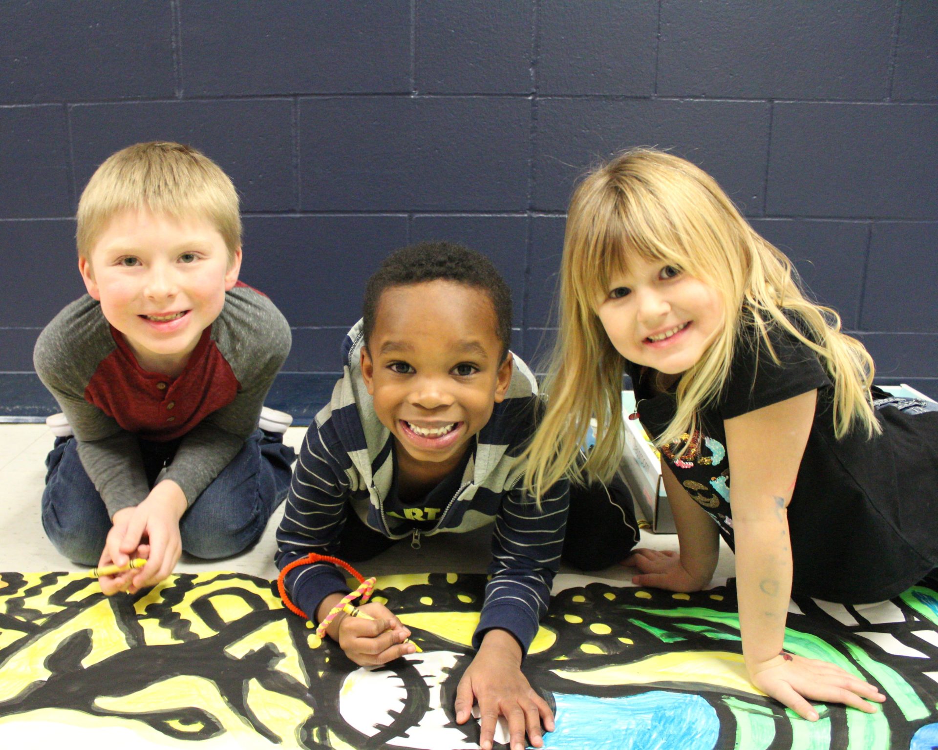 Three students smile as they color on the floor banner.