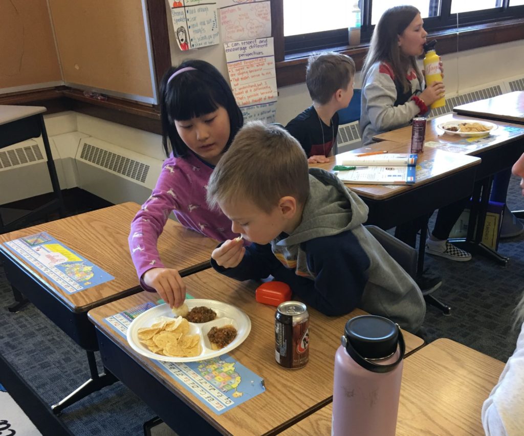 Two students sit at their desks trying salsa and chips.