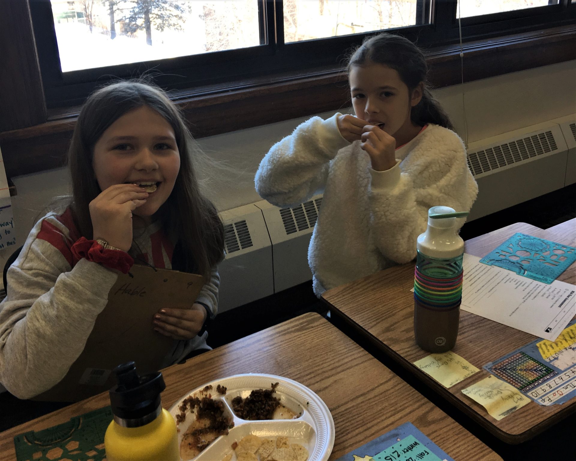 Two students sit at their desks trying salsa and chips.